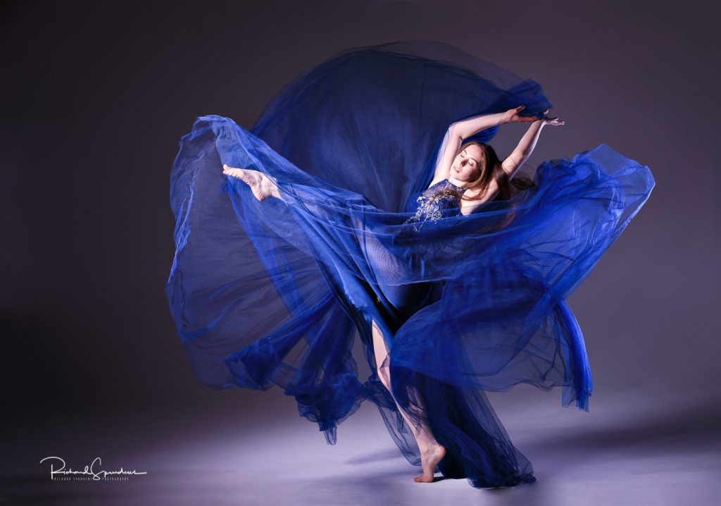 dance photographer - dance photography - colour image of the dancer wearign a large floaty blue dress kicking her right leg high out the the lhs of her with her arms reaching back behind her