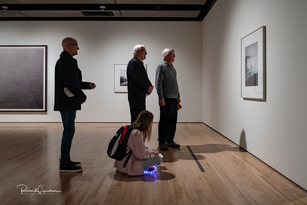 travel photography - travel photograher - colour image from a visit to the hayward gallery focusing on the visitors viewing the Gursky images in this one three man standing at one image on the floor a young student sits on the floor looking at the same image but her trainers are illuminated and this makes it different image