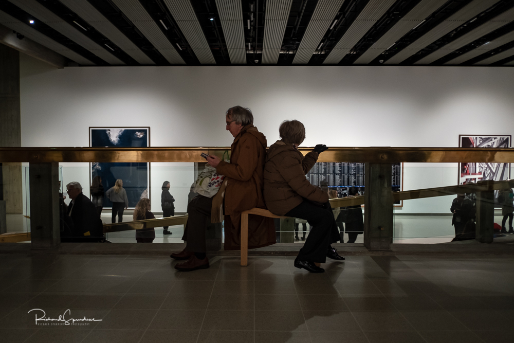 travel photography - travel photograher - colour image from a visit to the hayward gallery focusing on the visitors viewing the Gursky images in this image two visitors are sat back to back on a small bench he is reading his guide she is looking at other visitor below and behind her