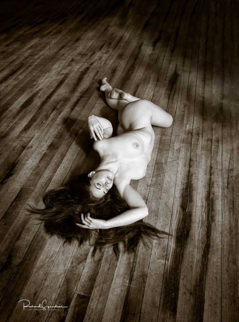 artistic nude photography - artistic nude photographer - toned monochrome image of artistic nude model elle beth making shapes on a wooden textured floor and showing the natural light patterns and