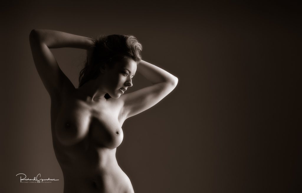 Fine Art Nude Photography - Fine Art Nude Photographer - Fine art nudes - toned monochrome image of rosa with her hands behind her head looking down towards the rhs of the image the lighting is highlighting her face and left breast the rest of her figure is in shadow