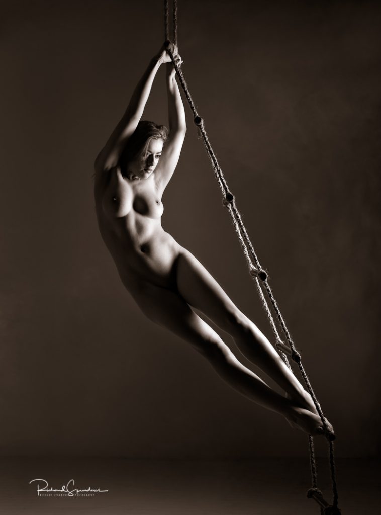 Fine Art Nude Photography - Fine Art Nude Photographer - toned monochrome image of rosa hanging away for a set of rope ladders her figure is arching away to the left hand side of the lighting which is coming in from the rhs