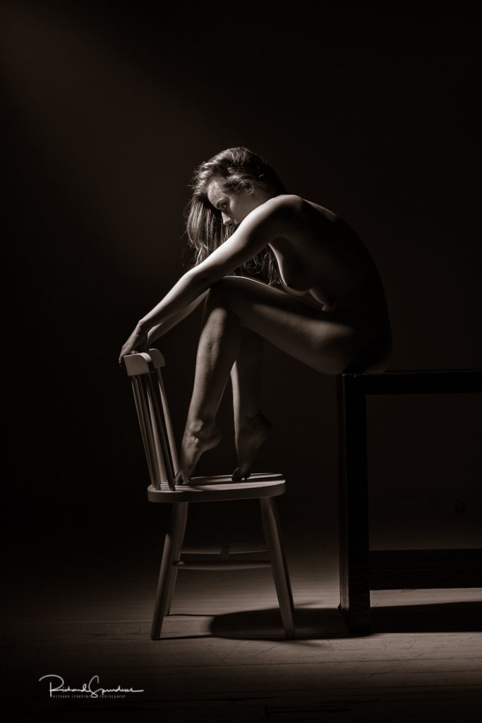Fine Art Nude Photography - Fine Art Nude Photographer - toned monochrome iamge of rosa she is sitting on a table to at the rhs of the image her feet are on a chair and her hands a resting on its back. the light is coming in from the lhs and is highlighting her arms arms and shoulder and hair the rest of her figure is in shadows