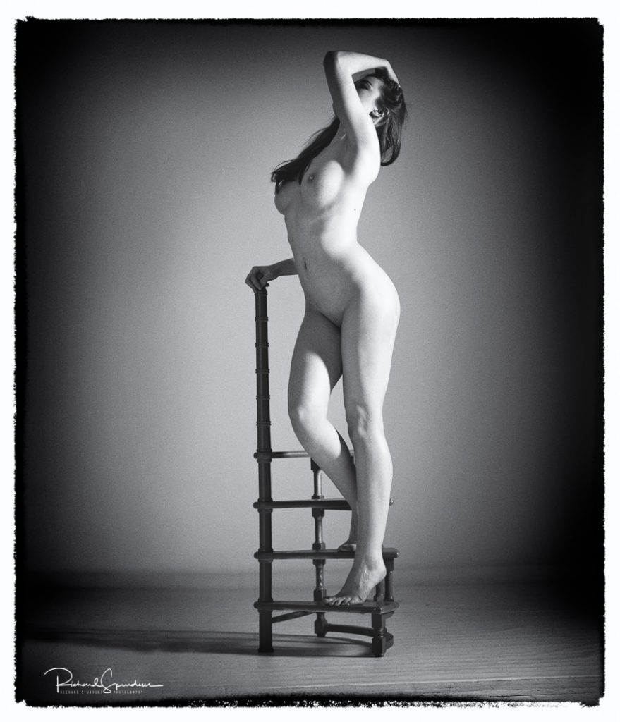 Fine Art Nude Photography - Fine Art Nude Photographer - monochrome image of rose standing on small set of library type circular steps she is reaching her nearest to the camera hand to the top of her head, the other is holding the step post.
