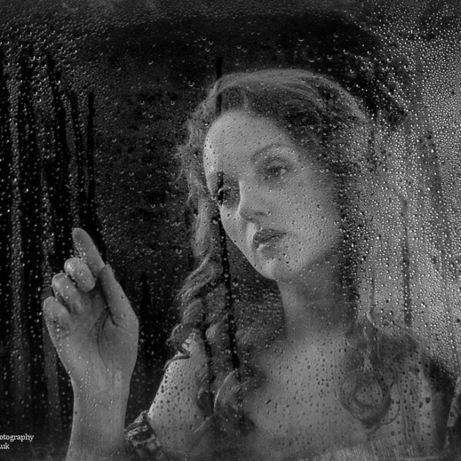 portrait photography - portrait photographer - monochrome image of a model standing behind a window with rain drops runing down the outside. she is reaching her finger to trace a rain drop down the window