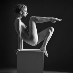 artistic nude photographer - artistic nude photography - monochrome image the model in using a white posing box she pushing up with her arms at the back of the box and her left foot to lift herself off the box her other leg is lifted and pointed at ninty degress towards the light. which is a single side light to the right hand side of the model.