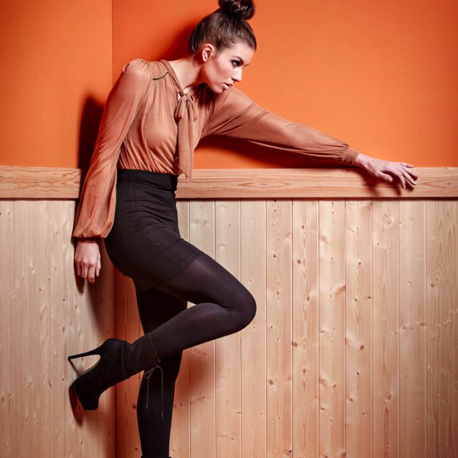 fashion photographer - fashion photography - colour image from a studio fashion shoot model weating back boots and tights with a black skirt and muted orange top. she is in the corner of an orange painted walled room with wood panelling from the floor to half way up the wall she is looking sideways along the wall