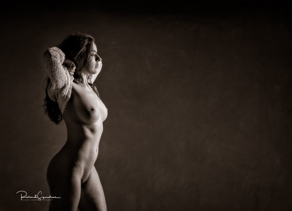 Fine Art Nude Photography - Fine Art Nude Photographer - a toned monochrome image of rose stood in profile with her arms behind her head she is on the lhs of the image and the lighting is coming in from the rhs and producing light and shadow across her figure.