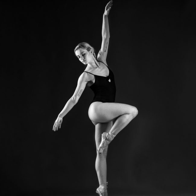 dance Photographer - dance photography - monochrome image of dance holding a raised leg on pointe pose with one arm reaching up above her head and the other down and out to the raised leg side. she is wearing a black leotard and pointe shoes