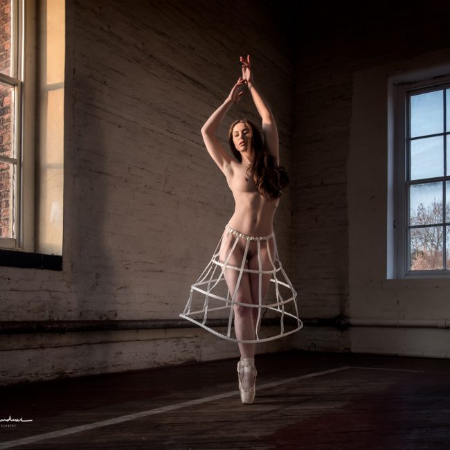 artistic nude photographer - artistic nude photography - colour image of model art nude model wear a crinoline cage dress pannier together with pointe shoes . she is on ponite and reaching her arms above her head there are two large windows one to her left the other to her right and the fading evening sun is lighting her body shapes