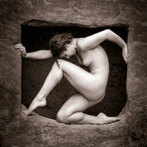 artistic nude photographer - artistic nude photography - a tonned monochrome image artistic nude model Micshkan she is posing in a rock opening, her arms are pushing at the sides and she is nearing on one leg and the other is pointed into the opposite corner. shot on location Guys Cliffe.