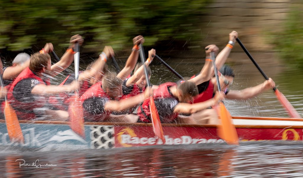 sport photography - sport photographer -colour image of dragon boat racing on the river aire at roberts park saltaire, a slow shutter speed was used together with panning to give a feel of the speed the on camera flash has frozen the water splash from the effort the paddlers are puttting in to power the boat foreard