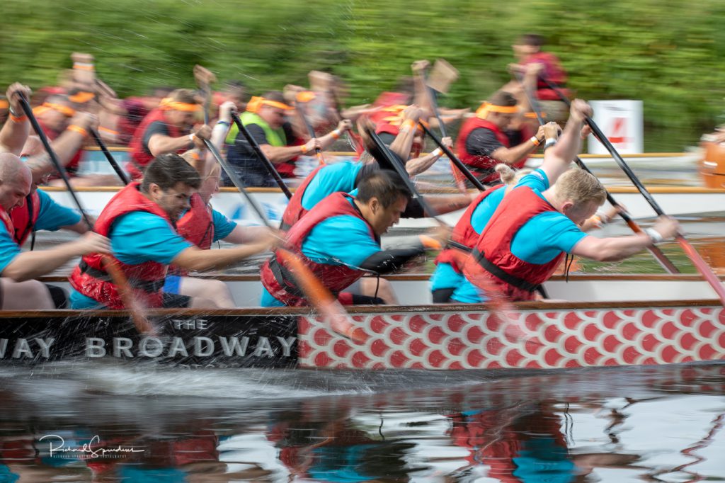 sport photography - sport photographer -colour image of dragon boat racing on the river aire at roberts park saltaire, a slow shutter speed was used together with panning to give a feel of the speed the colours the team in the front boat of this image are blue and red and stand out, as they paddle hard past me