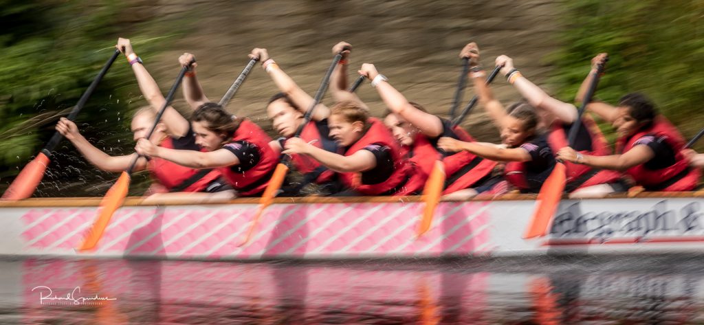 sport photography - sport photographer -colour image of dragon boat racing on the river aire at roberts park saltaire, a slow shutter speed was used together with panning to give a feel of the speed and in this image a feeling of really speed as all the paddles have the paddle at the top of the stroke and they arms are all up in the air