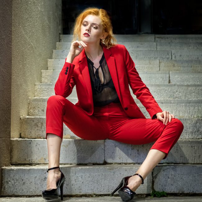 fashion photographer - fashion photography - colour image from a night shoot with model kate ri - she is wearing a red trouser suit she is sat on steps light by the light of a shop window, which is providing the light for the shot