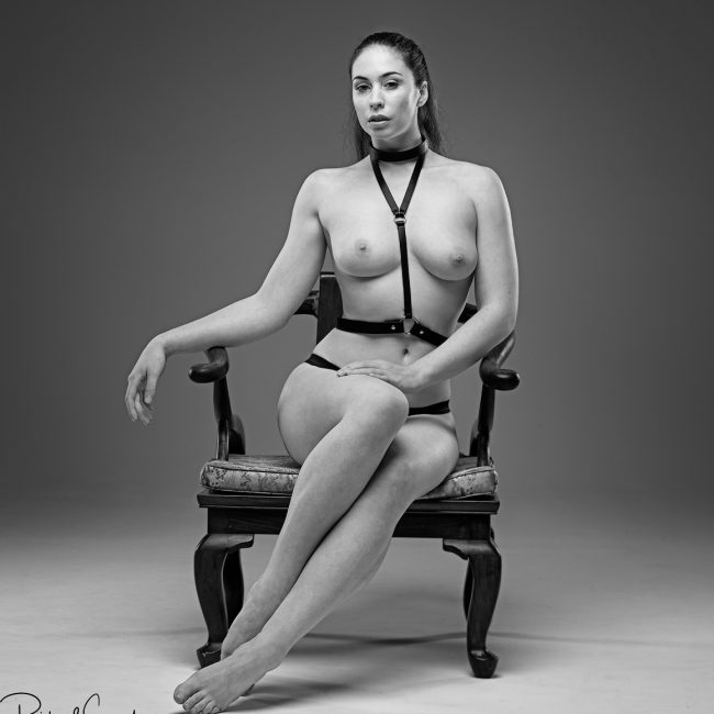 artistic nude photography - artistic nude photographer - monochrome image of model elle beth wearing a simple black leather harness and posing towards the camera with a relaxed look with her arm resting on the arm of the chair and on her forward knee