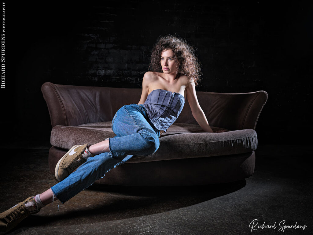 fashion photography fashion photographer - model sitting on the edge of a settee wearing blue demin jeans and demin top stretch her legs toward to lhs of the image