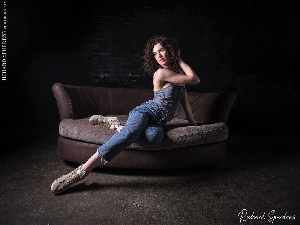 fashion photography fashion photographer - model poseing on the edge of a settee wearing blue demin jeans and demin top stretch her leg out.