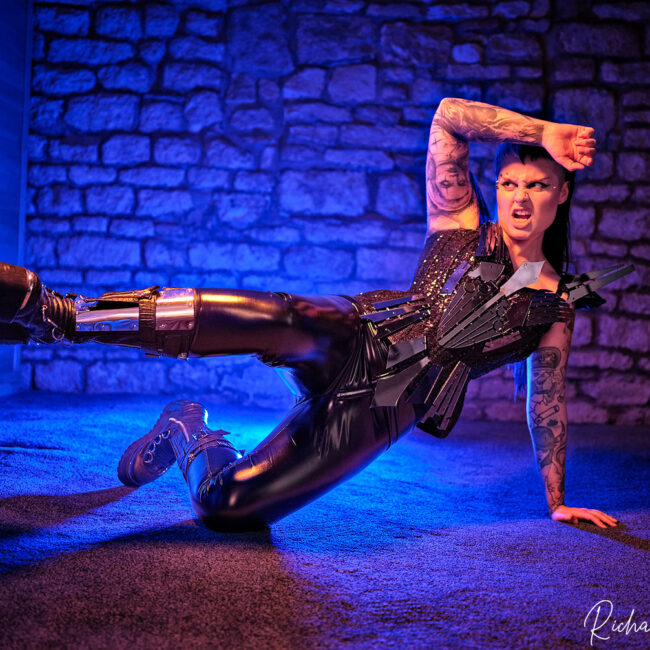 fashion photography fashion photographer - model wearing futuristic designes by bibian blue, black shiny trousers and big gothic boots is kicking out to her side from the floor .