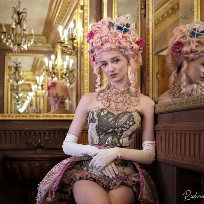 fashion photography fashion photographer - wearing colourful baroque era corset jewels and a pouf emblazoned with flowers and bird in a birdcage gives a elegent boudoir look.